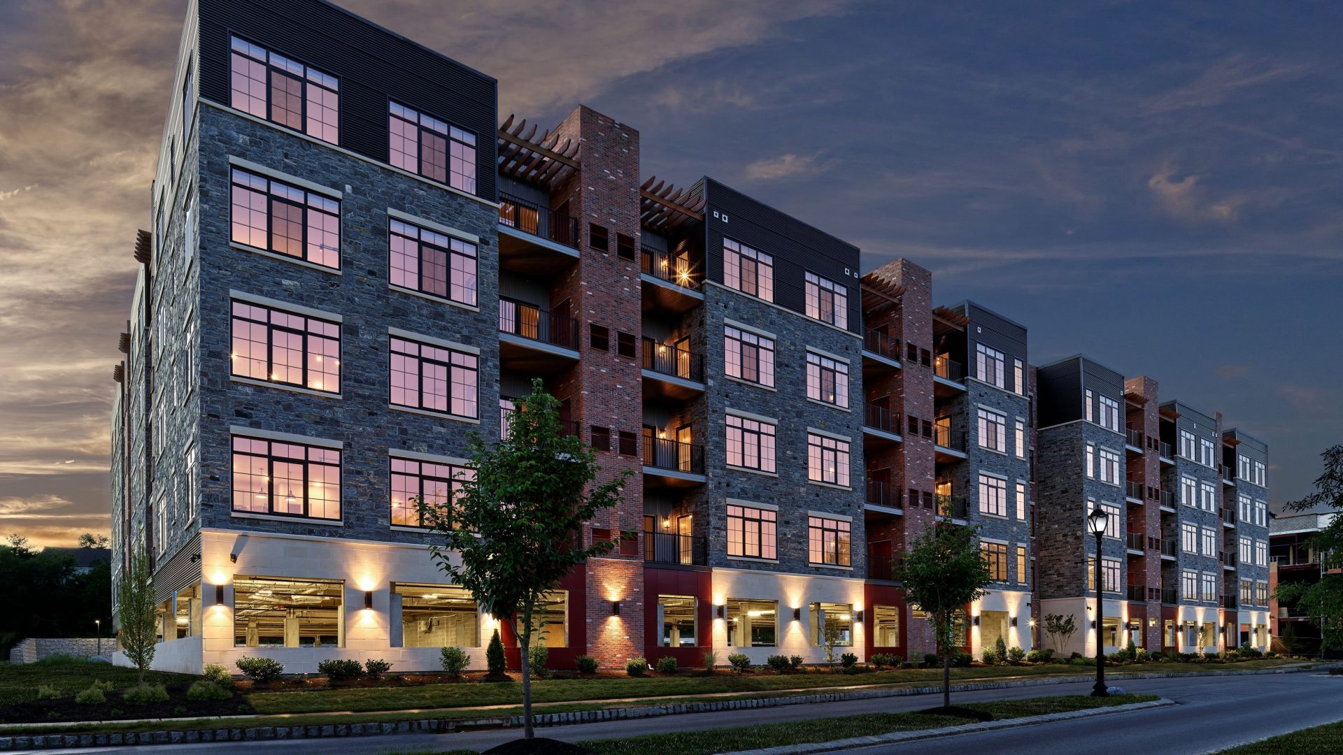 Multi-story stone and brick apartment building illuminated at dusk with landscaped entrance and street lighting