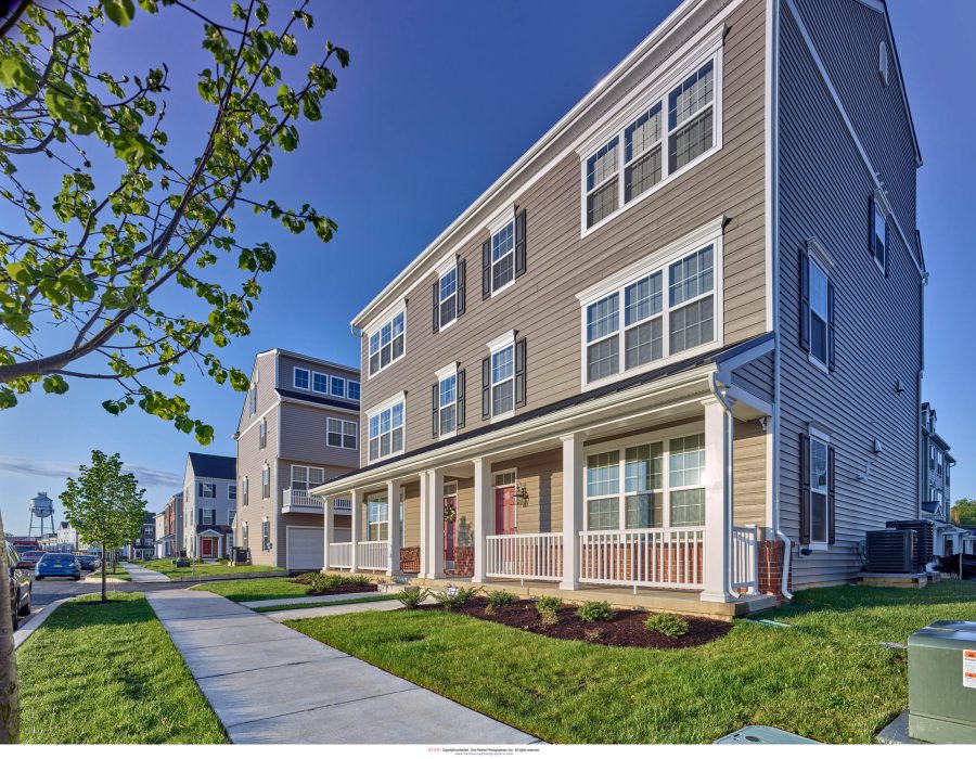 Three-story townhomes with tan siding, white trim, and a covered front porch along a residential street