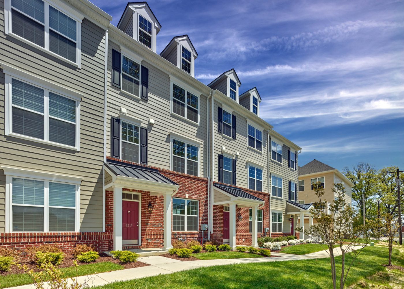 Three-story brick and gray-sided townhomes with dormer windows and red front doors along a landscaped walkway.