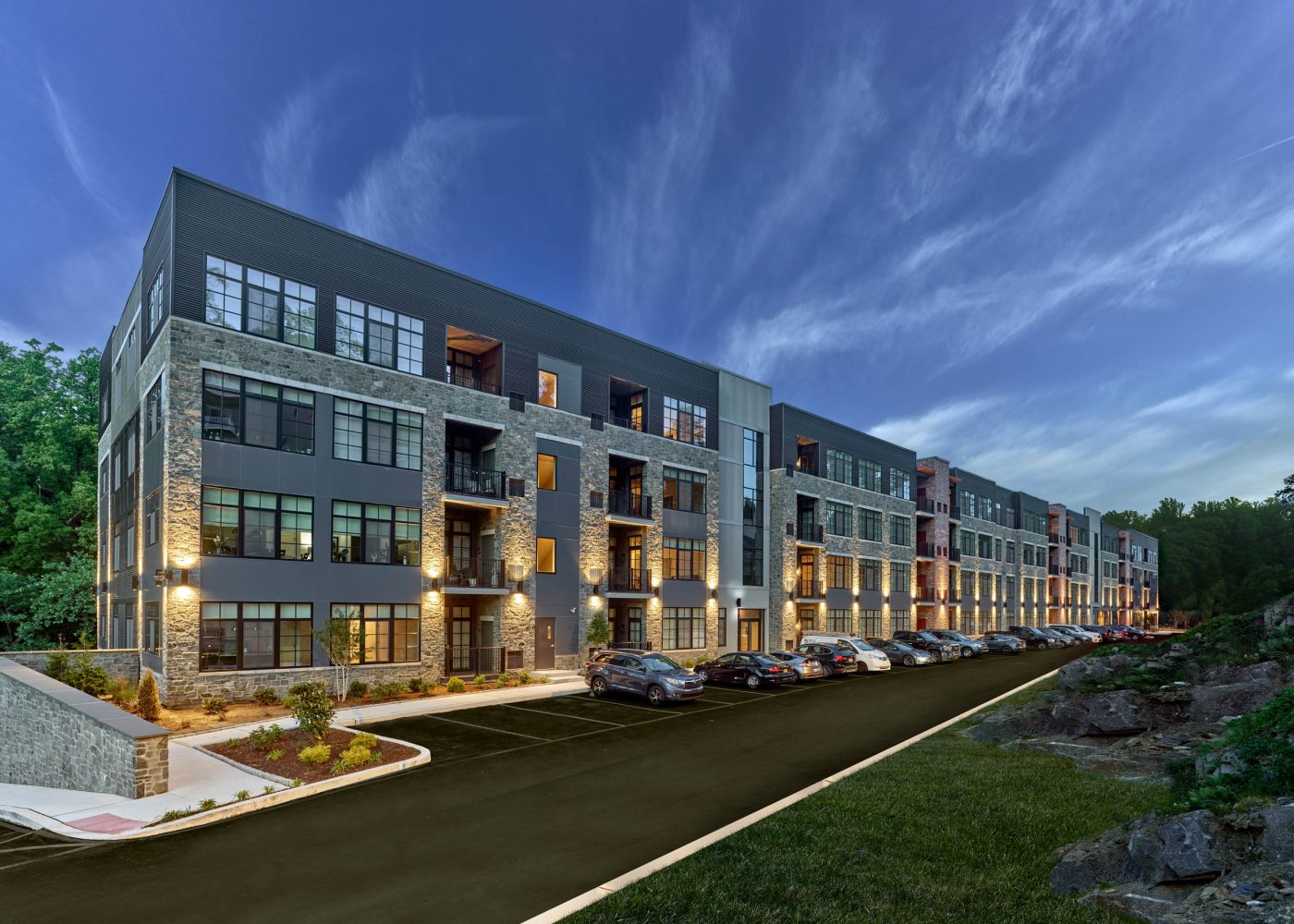 Multi-story stone and metal apartment building at dusk with illuminated windows and a parking lot in front.