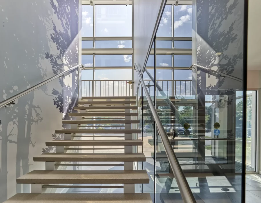 Open staircase with floating concrete steps and glass railings beside a tall window wall and tree-patterned walls