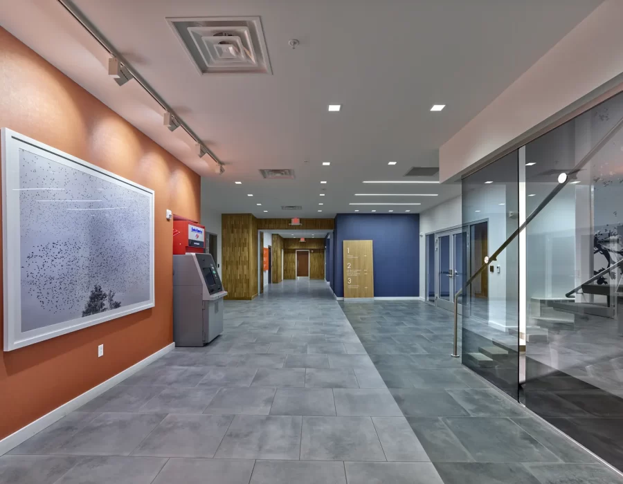Modern office lobby with orange accent wall, large artwork, ATM, blue wall signage, and glass stairwell