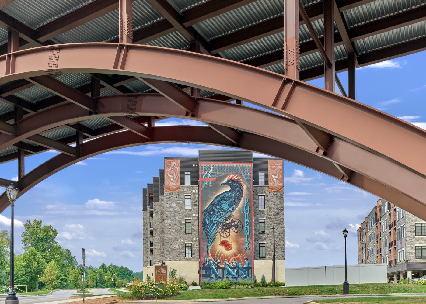 Steel canopy arches over The Phoenix apartment building featuring a large bird mural and leasing banners