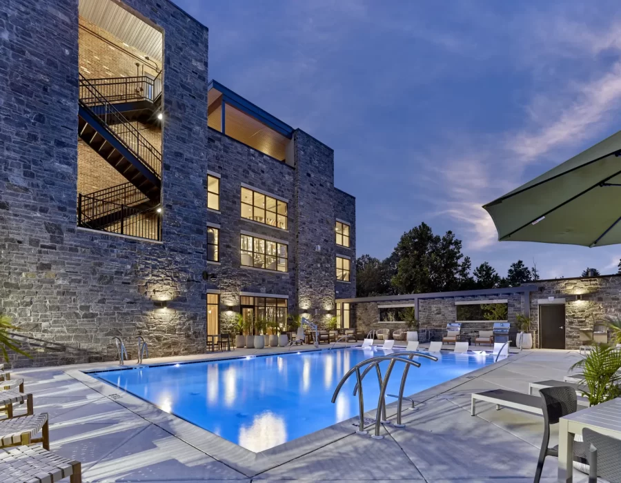 Stone-clad apartment building at dusk with glowing windows overlooking an outdoor pool and lounge area