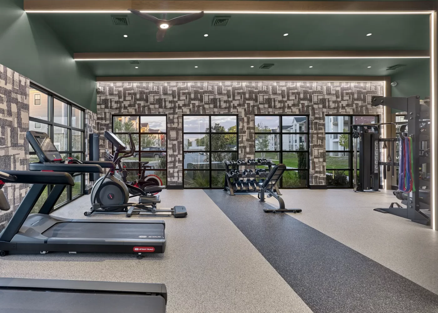Apartment fitness center with treadmills, elliptical machines, dumbbell rack, and cable weight station near large windows.