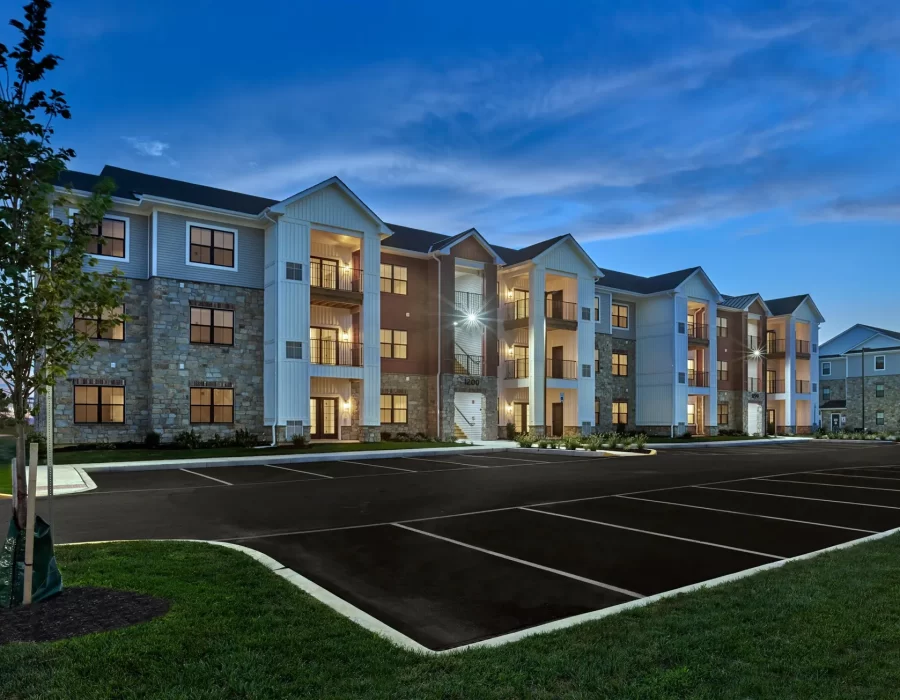 Three-story apartment building with stone and siding exterior illuminated at dusk, with parking lot and landscaping in front.