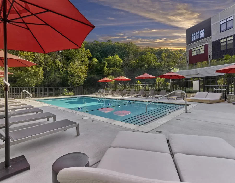 Red umbrellas shade lounge chairs beside an outdoor pool with trees and an apartment building in the background