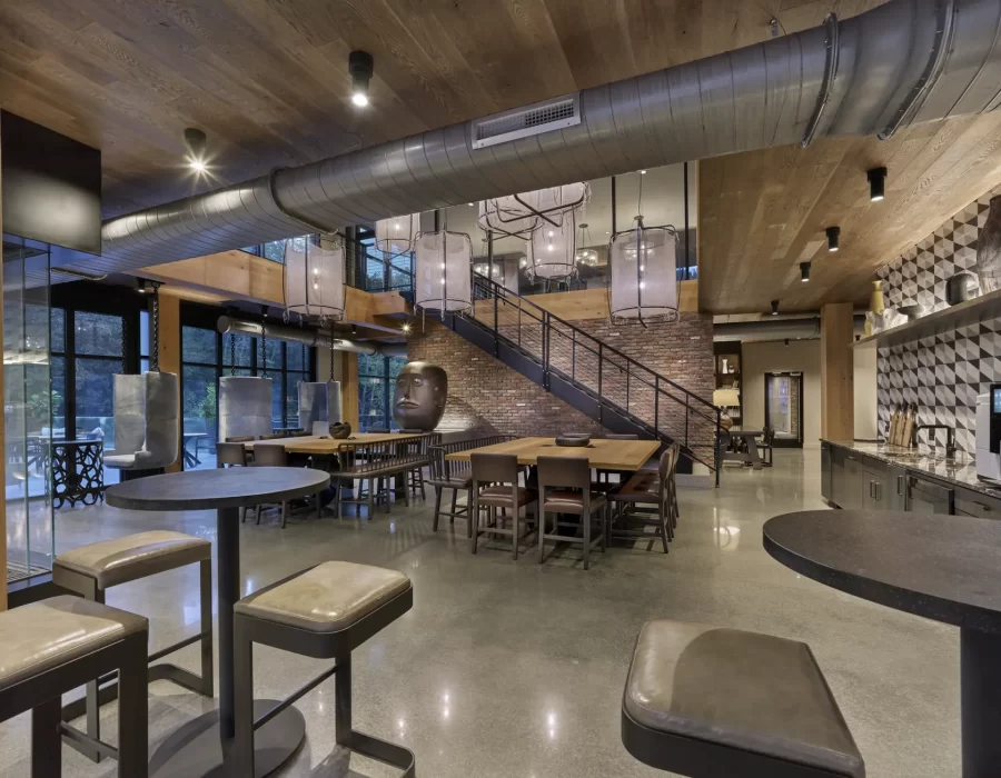 Industrial-style lounge with exposed ductwork, wooden tables, bar stools, and a staircase leading to a loft
