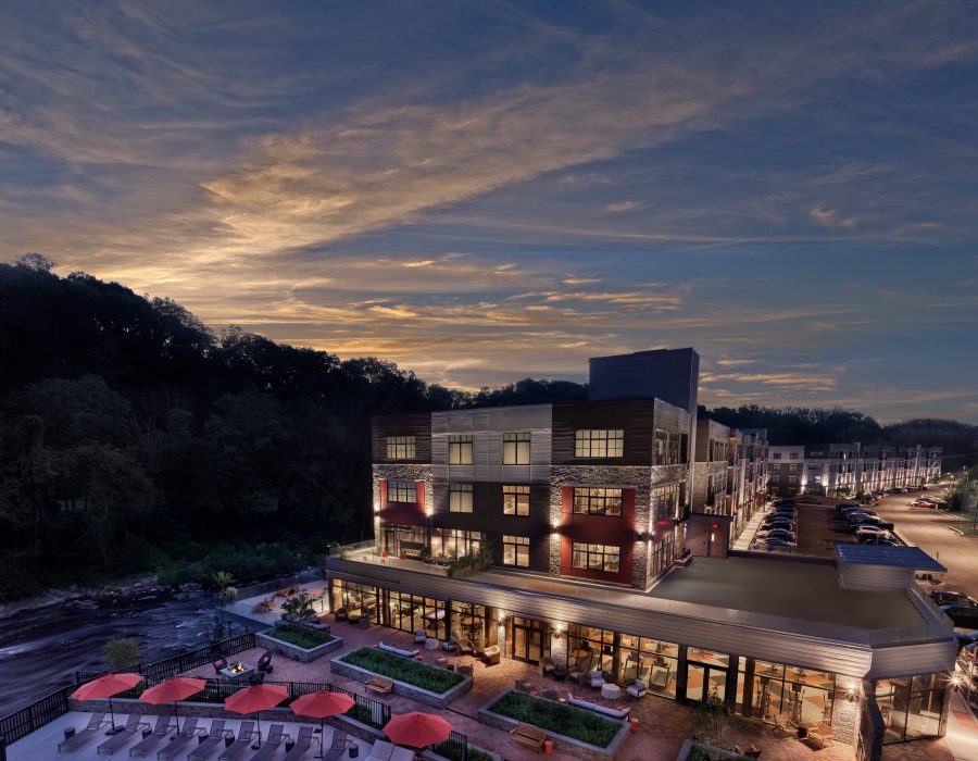 Riverside apartment building at dusk with lit windows, rooftop patio, and red umbrellas by the water