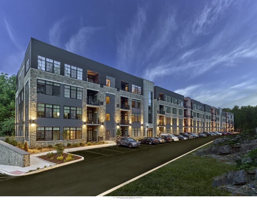 Multi-story apartment building with stone and metal facade, lit balconies, and a parking lot at dusk