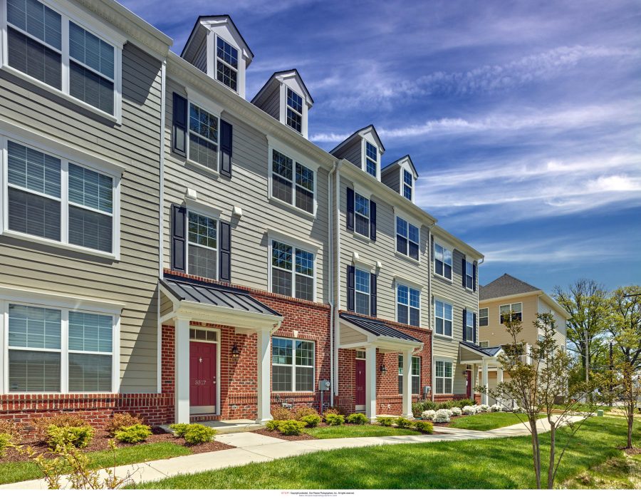 Row of three-story townhomes with gray siding, brick accents, red doors, and dormer windows under a blue sky