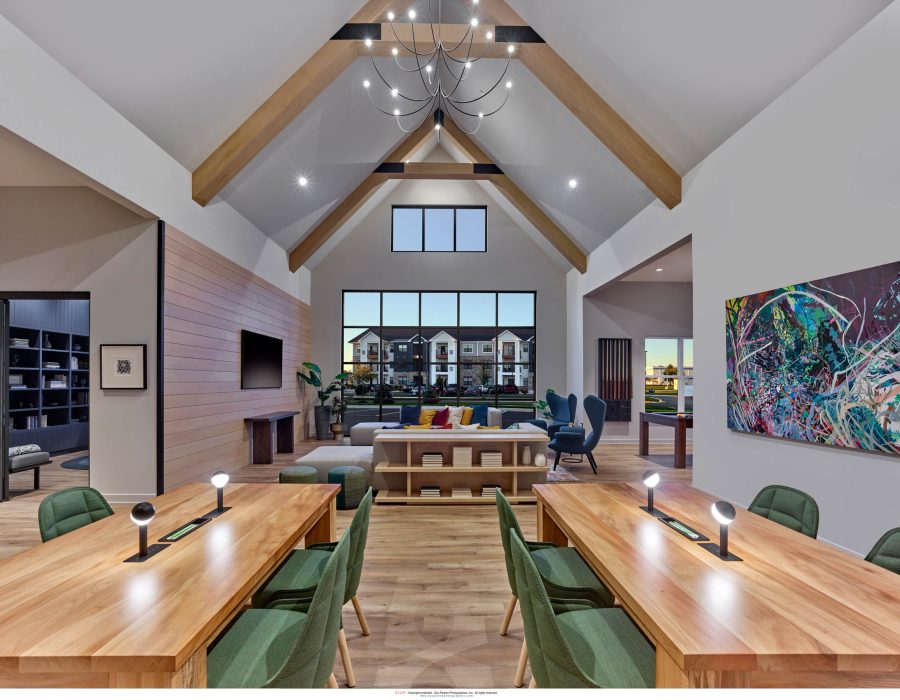 Vaulted clubhouse lounge with exposed wood beams, long work tables, green chairs, and a seating area beyond.