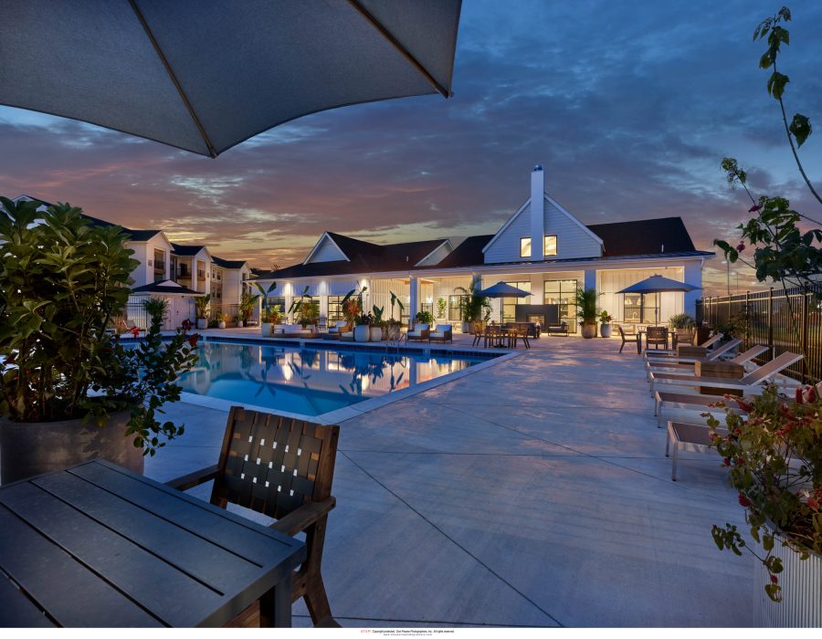 Apartment community pool and clubhouse lit up at dusk with lounge seating and umbrellas