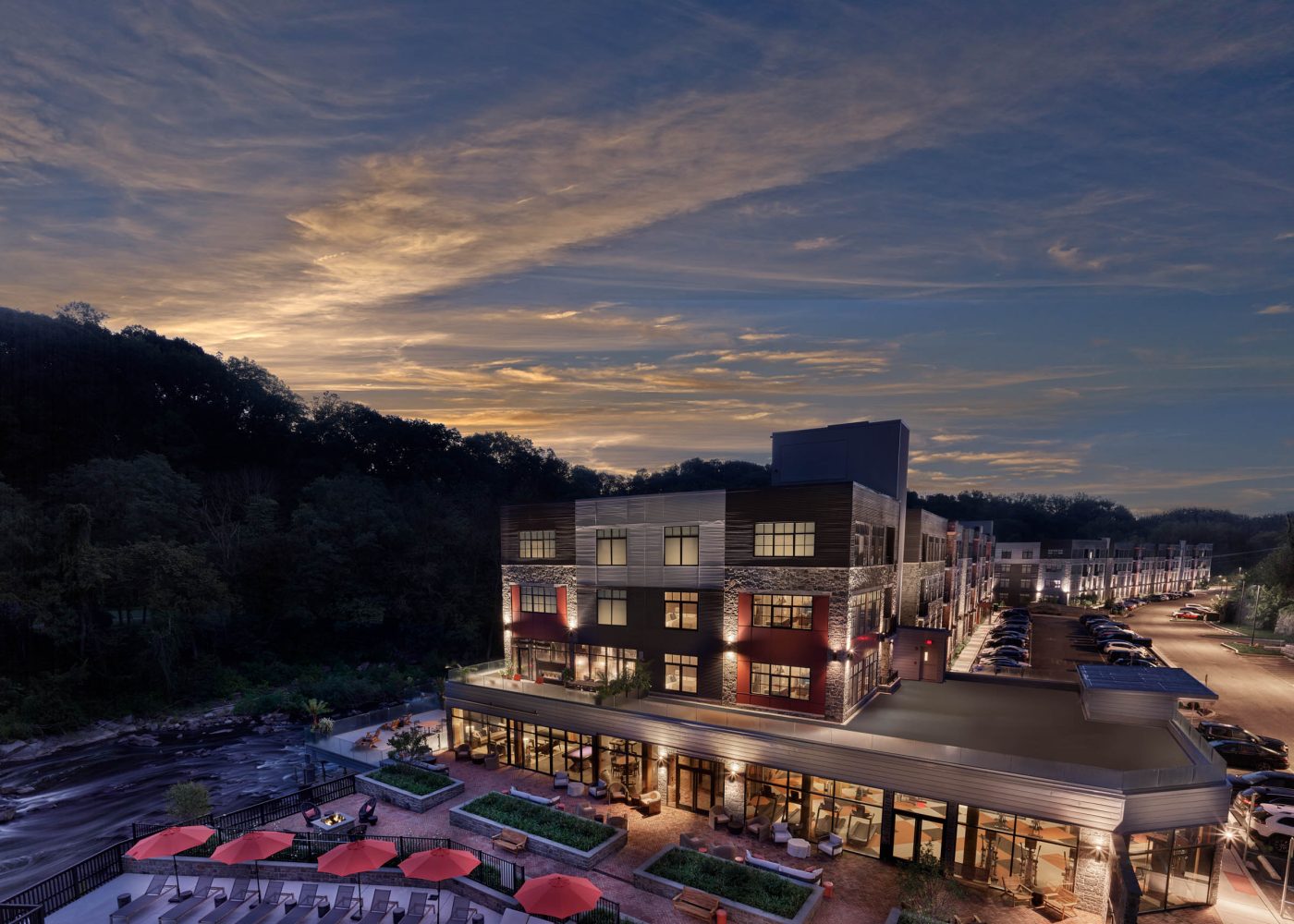 Multi-story apartment building at twilight with lit windows, riverside patio, pool loungers, and red umbrellas