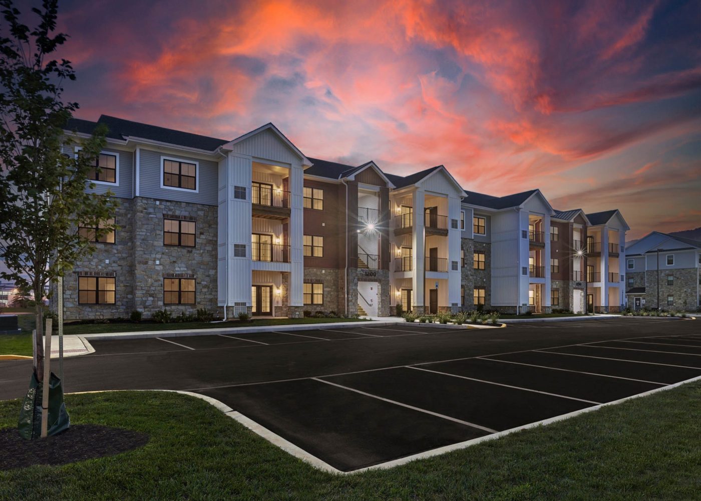 Three-story apartment building with stone and siding exterior, lit entrances, and a vivid sunset sky